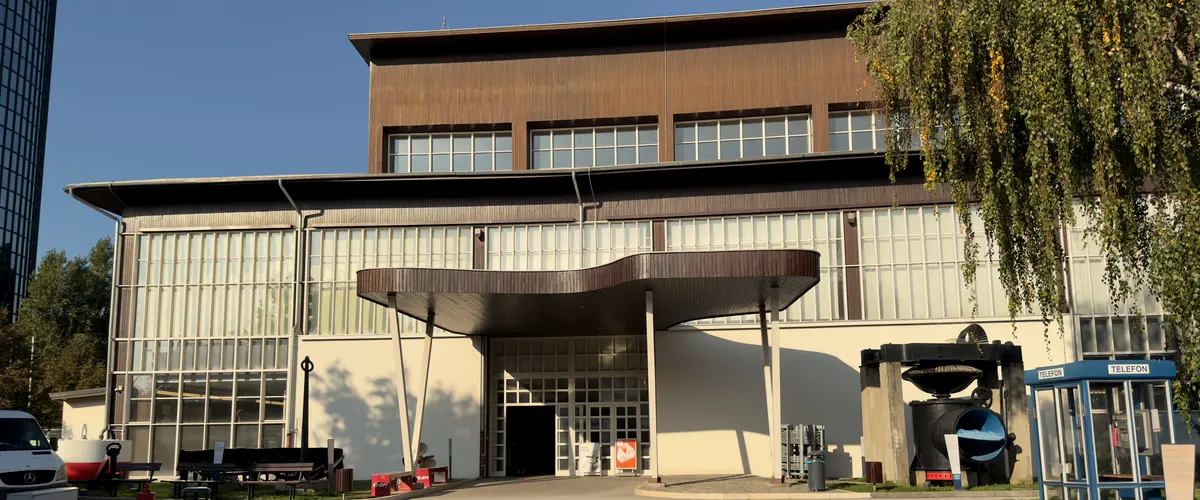 Der Eingang des Tehnički muzej Nikola Tesla in der Savska cesta 18: das geschwungene Holzvordach, die Glasfassade und rechts eine Turbine und eine Telefonzelle.